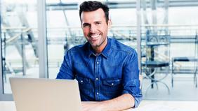 MBA IT-Management Lächelnder Mann in Freizeitbusiness-Kleidung sitzt vor einem Laptop in moderner Umgebung – symbolisiert den digitalen, ortsunabhängigen Zugang zum MBA IT-Management der FH Burgenland.