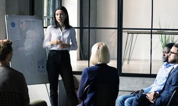 Junge Frau präsentiert vor einer kleinen Gruppe im Business-Setting; im Hintergrund ein Flipchart mit Diagrammen.