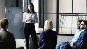 Junge Frau präsentiert vor einer kleinen Gruppe im Business-Setting; im Hintergrund ein Flipchart mit Diagrammen.