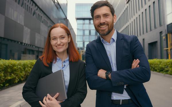 Flexibles Masterstudium an der Hochschule Burgenland: MBA, LL.M., MA (CE) und MSc (CE) Zwei junge Berufstätige stehen lächelnd vor einem modernen Bürogebäude. Sie symbolisieren das flexible Masterstudium an der Hochschule Burgenland.