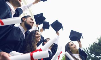 Eine Gruppe junger Absolventinnen und Absolventen in Talaren und mit Hüten feiert ihren Studienabschluss im Freien, die Hüte werden jubelnd in die Luft geworfen.