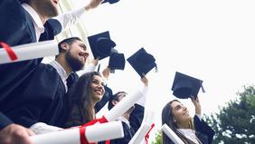 Eine Gruppe junger Absolventinnen und Absolventen in Talaren und mit Hüten feiert ihren Studienabschluss im Freien, die Hüte werden jubelnd in die Luft geworfen.