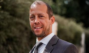 Porträt von Thomas Bravo im Anzug mit Krawatte, aufgenommen im Freien. Im Hintergrund sind unscharfe grüne Bäume und ein helles Gebäude erkennbar – Symbolbild für beruflichen Erfolg und persönliche Weiterentwicklung.