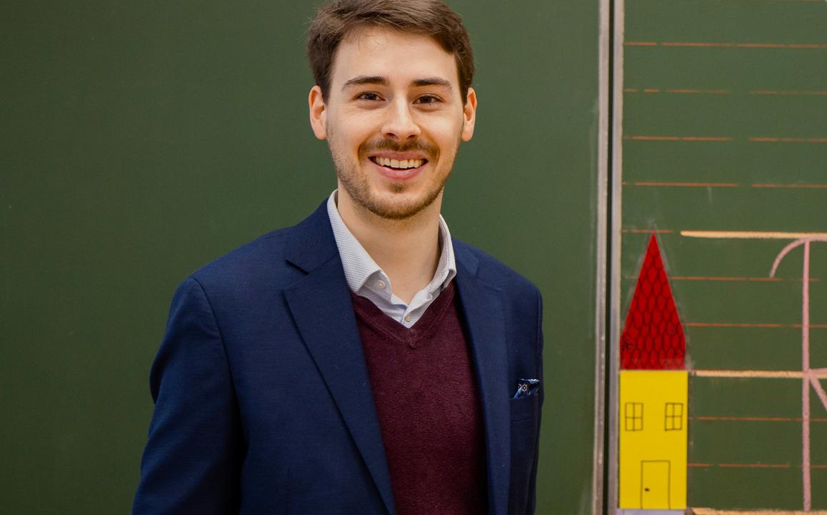 Constantin Haider steht lächelnd vor einer Tafel mit Zeichnungen und Diagrammen – symbolisiert akademische Expertise und praxisnahe Lehre.