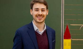 Constantin Haider steht lächelnd vor einer Schultafel – symbolisiert akademische Expertise und praxisnahe Lehre.