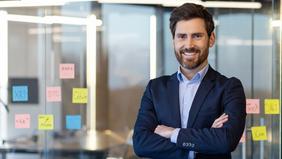 MBA Wirtschaftsinformatik Selbstbewusster junger Mann mit Vollbart und Anzug steht mit verschränkten Armen vor einer Glaswand mit bunten Post-its. Die Umgebung wirkt modern und innovativ – ideal passend zur praxisorientierten Weiterbildung im Bereich Wirtschaftsinformatik.