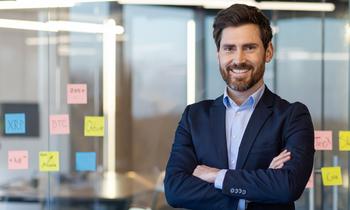 MBA Wirtschaftsinformatik Selbstbewusster junger Mann mit Vollbart und Anzug steht mit verschränkten Armen vor einer Glaswand mit bunten Post-its. Die Umgebung wirkt modern und innovativ – ideal passend zur praxisorientierten Weiterbildung im Bereich Wirtschaftsinformatik.