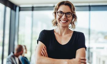 Junge Frau mit lockigen blonden Haaren, schwarzem T-Shirt und Brille steht mit verschränkten Armen lächelnd in einem modernen, lichtdurchfluteten Büro – Symbolbild für das MBA-Studium in Crossmediales Marketing, Event- und Kommunikationsmanagement an der Hochschule Burgenland.
