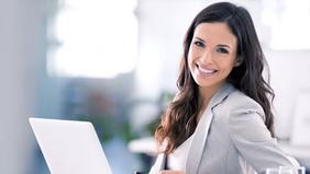 Junge Frau im Business-Outfit lächelt selbstbewusst in die Kamera, während sie an einem Laptop arbeitet – Symbolbild für den neuen MBA-Lehrgang Marketing und Kommunikationsmanagement der FH Burgenland.