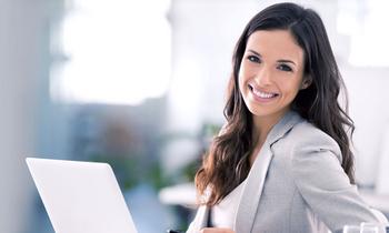 Junge Frau im Business-Outfit lächelt selbstbewusst in die Kamera, während sie an einem Laptop arbeitet – Symbolbild für den neuen MBA-Lehrgang Marketing und Kommunikationsmanagement der FH Burgenland.