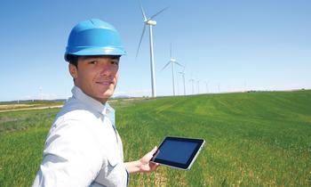Mann mit Schutzhelm und Tablet steht auf einem grünen Feld vor mehreren Windrädern und prüft intelligente Energiesysteme im Rahmen eines nachhaltigen Hochschullehrgangs.