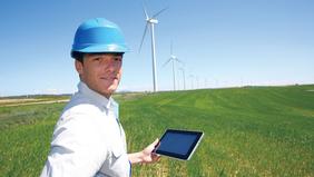 Mann mit Schutzhelm und Tablet steht auf einem grünen Feld vor mehreren Windrädern und prüft intelligente Energiesysteme im Rahmen eines nachhaltigen Hochschullehrgangs.