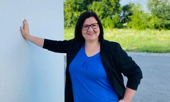 Johanna Prodinger, eine Studentin der FH Burgenland, steht lächelnd vor einer hellen Wand im Freien. Sie trägt ein blaues Oberteil mit dunklem Blazer und blickt selbstbewusst in die Kamera.