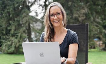 Elke Egi sitzt entspannt im Garten an einem Tisch und arbeitet an einem silbernen Laptop – ein Symbol für Flexibilität und Selbstbestimmung im Fernstudium.