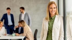 Junge Frau mit blondem Haar in grünem Kleid und hellem Blazer steht lächelnd vor einer Glaswand in einem modernen Büro. Im Hintergrund arbeiten mehrere Personen im Business-Outfit in einer Besprechung. Das Bild vermittelt Professionalität, Führungskompetenz und berufliche Weiterentwicklung – passend zum MBA Personalführung der FH Burgenland.