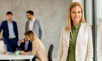 Junge Frau mit blondem Haar in grünem Kleid und hellem Blazer steht lächelnd vor einer Glaswand in einem modernen Büro. Im Hintergrund arbeiten mehrere Personen im Business-Outfit in einer Besprechung. Das Bild vermittelt Professionalität, Führungskompetenz und berufliche Weiterentwicklung – passend zum MBA Personalführung der FH Burgenland.