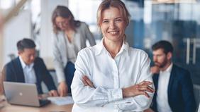 MBA Change Management und Leadership Selbstbewusste Frau in weißer Bluse steht mit verschränkten Armen im Vordergrund eines modernen Büros. Im Hintergrund arbeiten mehrere Personen konzentriert. Das Bild symbolisiert Führungskompetenz und berufliche Weiterentwicklung – passend zum MBA Change Management und Leadership der FH Burgenland.
