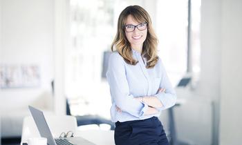 Junge, lächelnde Frau mit Brille und verschränkten Armen steht selbstbewusst vor einem Schreibtisch mit Laptop – symbolisiert modernes, flexibles Studieren im Bereich Marketingkommunikation an der FH Burgenland.