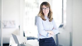Junge, lächelnde Frau mit Brille und verschränkten Armen steht selbstbewusst vor einem Schreibtisch mit Laptop – symbolisiert modernes, flexibles Studieren im Bereich Marketingkommunikation an der FH Burgenland.