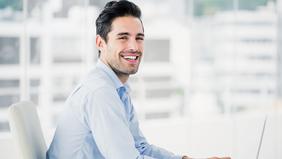 Lächelnder junger Mann in Business-Outfit sitzt vor einem Laptop in heller, moderner Umgebung – symbolisiert berufliche Weiterentwicklung im MBA Marketing, Event- und Kommunikationsmanagement der FH Burgenland.