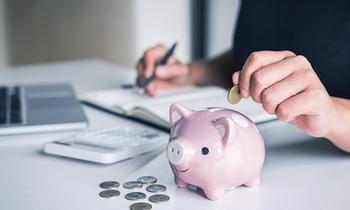Hand wirft Münze in rosa Sparschwein auf Schreibtisch, daneben Laptop und Notizbuch – Symbolbild für finanzielle Förderung und Weiterbildung.