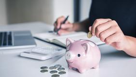 Hand wirft Münze in rosa Sparschwein auf Schreibtisch, daneben Laptop und Notizbuch – Symbolbild für finanzielle Förderung und Weiterbildung.