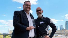 Zwei Männer bei sonnigem Wetter auf einer Dachterrasse, einer hält eine Glas-Trophäe in der Hand – Auszeichnung im Rahmen des BPUG-Kongresses für herausragende Abschlussarbeiten im Projektmanagement.