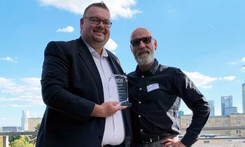 Zwei Männer bei sonnigem Wetter auf einer Dachterrasse, einer hält eine Glas-Trophäe in der Hand – Auszeichnung im Rahmen des BPUG-Kongresses für herausragende Abschlussarbeiten im Projektmanagement.