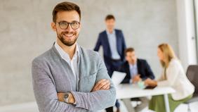 Selbstbewusster junger Mann mit Brille und grauem Sakko steht mit verschränkten Armen in einem modernen Büro. Im Hintergrund arbeiten mehrere Personen in Business-Kleidung an einem Tisch. Das Bild symbolisiert Professionalität und Karriereambitionen – ideal für den MBA Brand Management an der FH Burgenland.