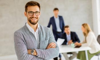 Selbstbewusster junger Mann mit Brille und grauem Sakko steht mit verschränkten Armen in einem modernen Büro. Im Hintergrund arbeiten mehrere Personen in Business-Kleidung an einem Tisch. Das Bild symbolisiert Professionalität und Karriereambitionen – ideal für den MBA Brand Management an der FH Burgenland.