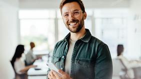 Diploma of Advanced Studies in Projektmanagement und New Leadership Ein junger, lächelnder Mann mit Tablet in der Hand steht selbstbewusst in einem modernen Büro – Symbolbild für moderne Führung und Projektmanagement im Rahmen des DAS Programms der Hochschule Burgenland.