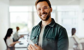 Ein junger, lächelnder Mann mit Tablet in der Hand steht selbstbewusst in einem modernen Büro – Symbolbild für moderne Führung und Projektmanagement im Rahmen des DAS Programms der Hochschule Burgenland.