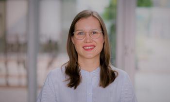 Porträt einer lächelnden Frau mit Brille und langen Haaren, im Hintergrund ein heller, unscharfer Flur mit Glasfronten.