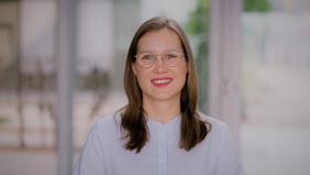 Porträt einer lächelnden Frau mit Brille und langen Haaren, im Hintergrund ein heller, unscharfer Flur mit Glasfronten.