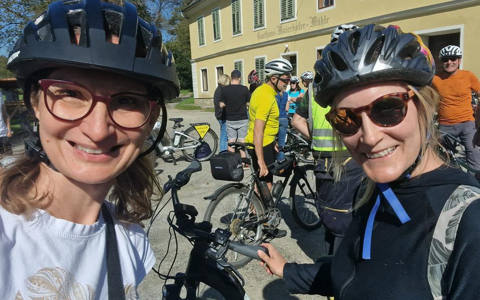 Zwei fröhliche Mitarbeiterinnen der FH Burgenland Weiterbildung mit Fahrradhelmen beim Start des Betriebsausflugs, im Hintergrund eine Gruppe von Kolleginnen und Kollegen mit Fahrrädern.