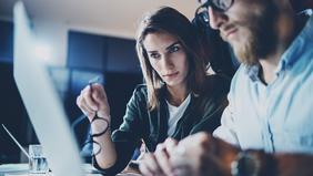 Zwei konzentriert wirkende Personen im Business-Outfit besprechen Unterlagen vor einem Laptop in einem modernen Büro – Symbolbild für den MBA-Lehrgang Compliance & Risikomanagement.