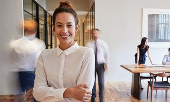 DAS Change Management, Resilienz und Stressmanagement Junge Frau mit zusammengebundenen Haaren in weißer Bluse steht lächelnd mit verschränkten Armen in einem modernen Büroflur. Im Hintergrund gehen und arbeiten mehrere Personen. Das Bild vermittelt Dynamik, Selbstsicherheit und den Fokus auf Resilienz und Stressmanagement im beruflichen Umfeld.