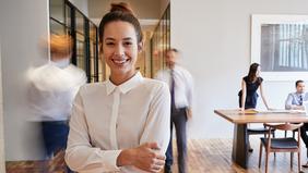 DAS Change Management, Resilienz und Stressmanagement Junge Frau mit zusammengebundenen Haaren in weißer Bluse steht lächelnd mit verschränkten Armen in einem modernen Büroflur. Im Hintergrund gehen und arbeiten mehrere Personen. Das Bild vermittelt Dynamik, Selbstsicherheit und den Fokus auf Resilienz und Stressmanagement im beruflichen Umfeld.