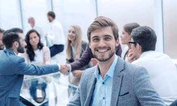 Fröhlicher junger Mann in Businesskleidung sitzt in einem modernen Konferenzraum, im Hintergrund ein Handschlag zwischen zwei Kolleg*innen – symbolisiert erfolgreiche Zusammenarbeit im Projekt- und Prozessmanagement.