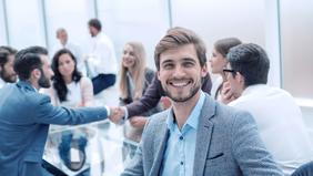 Fröhlicher junger Mann in Businesskleidung sitzt in einem modernen Konferenzraum, im Hintergrund ein Handschlag zwischen zwei Kolleg*innen – symbolisiert erfolgreiche Zusammenarbeit im Projekt- und Prozessmanagement.