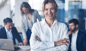 Selbstbewusste Geschäftsfrau in weißer Bluse steht mit verschränkten Armen im Vordergrund, im Hintergrund ein Team bei der Arbeit – symbolisiert Führungskompetenz und digitale Vertriebsstrategien im MBA Digital Sales und Vertriebsmanagement der FH Burgenland.