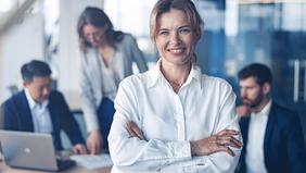 Selbstbewusste Geschäftsfrau in weißer Bluse steht mit verschränkten Armen im Vordergrund, im Hintergrund ein Team bei der Arbeit – symbolisiert Führungskompetenz und digitale Vertriebsstrategien im MBA Digital Sales und Vertriebsmanagement der FH Burgenland.