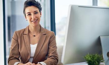 Fernstudium Digitales Bildungsmanagement Junge Frau im Business-Outfit sitzt lächelnd am Schreibtisch vor einem Computer – symbolisiert strategische Kompetenz und digitale Führung im Bildungsmanagement.
