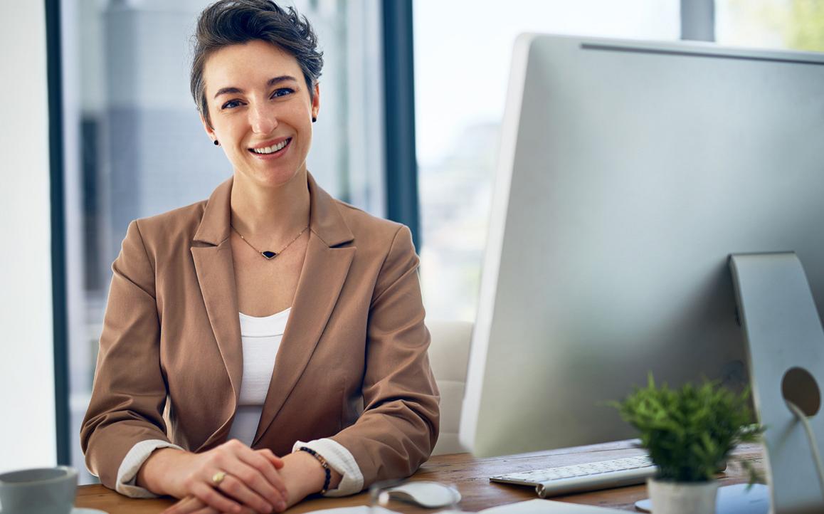 Junge Frau im Business-Outfit sitzt lächelnd am Schreibtisch vor einem Computer – symbolisiert strategische Kompetenz und digitale Führung im Bildungsmanagement.