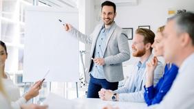 Junger Mann präsentiert motiviert vor einem Whiteboard einem aufmerksamen Team – Sinnbild für innovative Führung, modernes Leadership und interaktive Wissensvermittlung