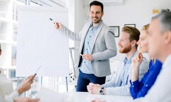 Junger Mann präsentiert motiviert vor einem Whiteboard einem aufmerksamen Team – Sinnbild für innovative Führung, modernes Leadership und interaktive Wissensvermittlung