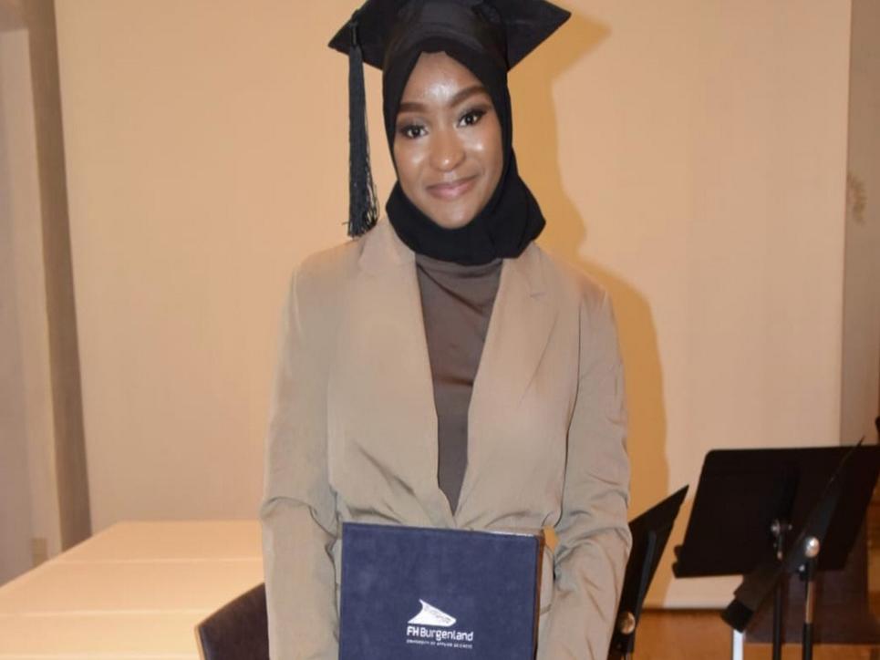 Safinatou Bella Bah mit Abschlusskappe und Diplommappe der FH Burgenland in den Händen, stolz lächelnd.