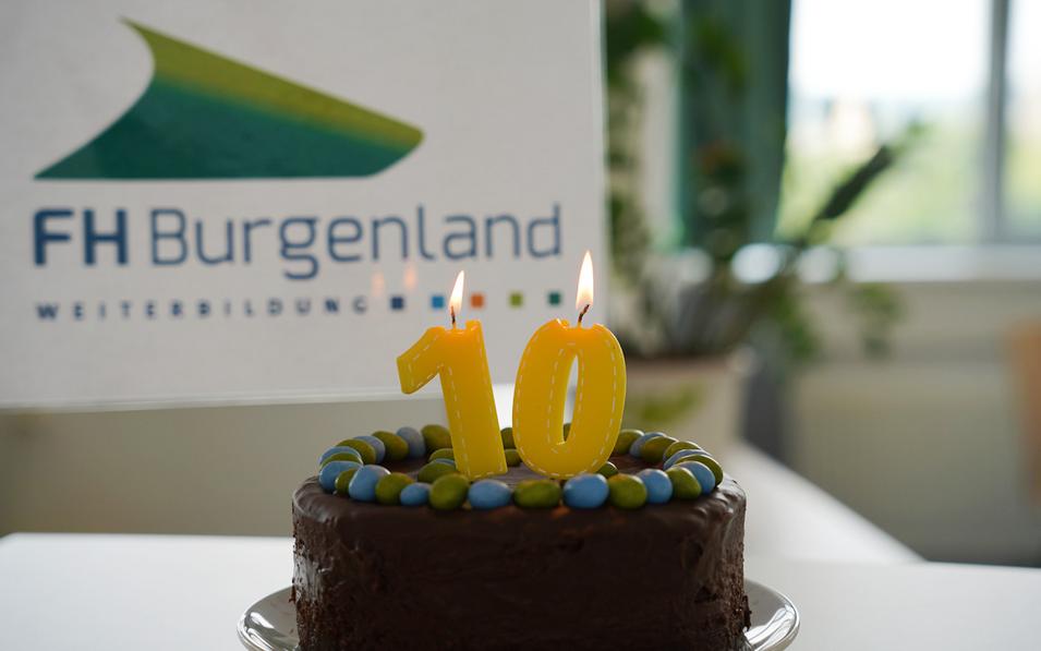 Schokoladentorte mit bunter Verzierung und einer Kerze in Form der Zahl 10 vor dem Logo der FH Burgenland Weiterbildung – Symbol für das 10-jährige Jubiläum.