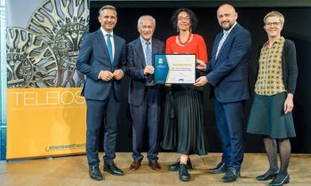 Gruppenfoto bei Preisverleihung: Fünf Personen stehen auf einer Bühne, einige davon halten eine Urkunde. Im Hintergrund ist ein dekoratives Element mit dem Schriftzug ‚TELEIOS‘ zu sehen. Die Stimmung ist feierlich, alle lächeln – passend zur Auszeichnung als EAN-Ausbildungseinrichtung.