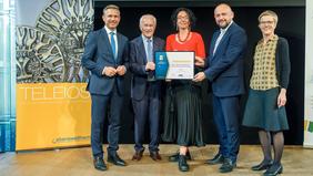 Gruppenfoto bei Preisverleihung: Fünf Personen stehen auf einer Bühne, einige davon halten eine Urkunde. Im Hintergrund ist ein dekoratives Element mit dem Schriftzug ‚TELEIOS‘ zu sehen. Die Stimmung ist feierlich, alle lächeln – passend zur Auszeichnung als EAN-Ausbildungseinrichtung.